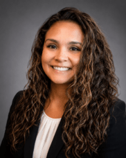 Professional headshot of a smiling woman with long, curly brown hair, wearing a black blazer and white blouse, against a gray studio backdrop.