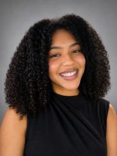 Portrait of a smiling woman with curly hair wearing a black sleeveless top, posing against a grey backdrop.