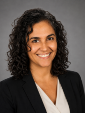 Professional head-and-shoulders portrait of a smiling woman with dark, curly hair in a black blazer and white blouse, against a neutral gray background.