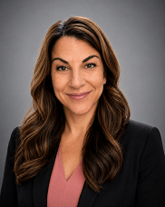 Professional headshot: smiling woman with brown hair, wearing a black blazer and pink blouse, gray background.