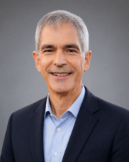 Smiling gray-haired man in a navy suit and light blue shirt, posing for a professional headshot against a neutral gray background.