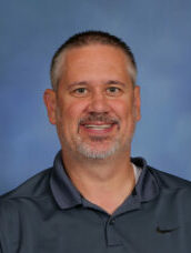 Smiling middle-aged man with short gray hair and beard, in a dark polo shirt, posing for a portrait against a blue background.