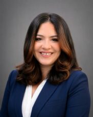 Professional headshot of a smiling woman in a navy blazer against a gray background, suitable for LinkedIn or corporate profiles.
