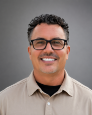 Head-and-shoulders portrait of a smiling man with black rectangular glasses and a beige shirt on a gray background.